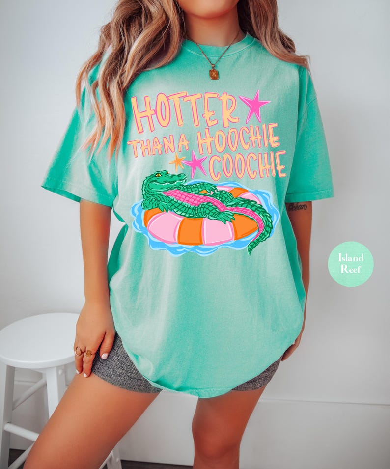 Puede incluir: Camiseta turquesa con el texto "Hotter Than a Hoochie Coochie" en rosa y naranja. La camiseta presenta un gr&aacute;fico de un caim&aacute;n verde sobre un flotador rosa y naranja. La camiseta tiene un ajuste holgado y c&oacute;modo.