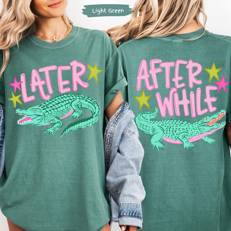 May include: Light green t-shirt with the words "LATER" and "AFTER WHILE" in pink, with a turquoise alligator graphic. The design includes pink and yellow stars. The shirt is a relaxed fit.