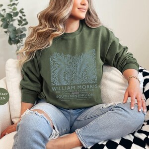 Puede incluir: Sudadera verde oliva con un dise&ntilde;o floral y el texto "WILLIAM MORRIS COTTON PRINTS EXHIBITION". La sudadera se combina con vaqueros azul claro. El estilo es informal y c&oacute;modo.
