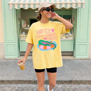 Puede incluir: Camiseta amarilla con el texto "Hotter Than a Hoochie Coochie" y un gr&aacute;fico de un caim&aacute;n en una colchoneta. La camiseta se usa con pantalones cortos negros y una gorra de b&eacute;isbol. La persona sostiene una bebida.
