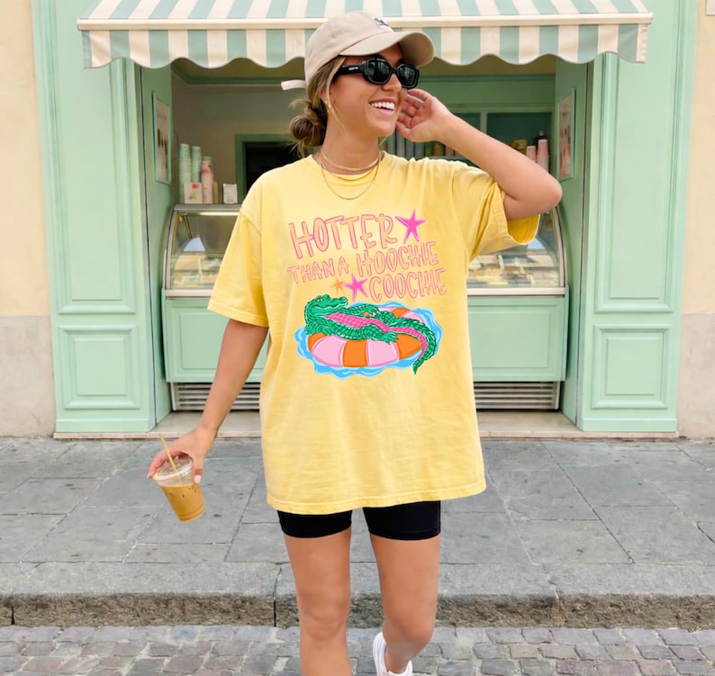 Puede incluir: Camiseta amarilla con el texto "Hotter Than a Hoochie Coochie" y un gr&aacute;fico de un caim&aacute;n en una colchoneta. La camiseta se usa con pantalones cortos negros y una gorra de b&eacute;isbol. La persona sostiene una bebida.