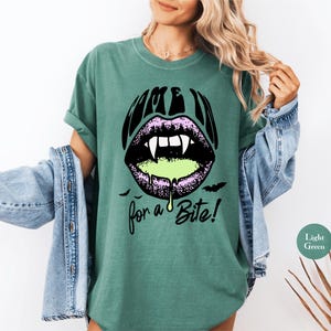 May include: A dark green t-shirt featuring a graphic of purple lips with white vampire fangs and dripping light green liquid. The text above the lips reads "COME IN" and the text below reads "for a Bite!" with small black bat silhouettes.