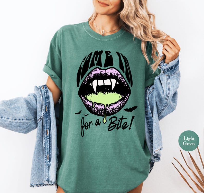 May include: A dark green t-shirt featuring a graphic of purple lips with white vampire fangs and dripping light green liquid. The text above the lips reads "COME IN" and the text below reads "for a Bite!" with small black bat silhouettes.