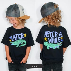 Puede incluir: Camisetas negras con un dise&ntilde;o de cocodrilo de dibujos animados. Una camiseta dice "LATER" con una estrella azul y amarilla, y la otra dice "AFTER A WHILE" con una estrella azul y amarilla. El ni&ntilde;o lleva una gorra de camionero.