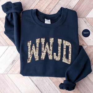 May include: Navy blue sweatshirt with the letters "WWJD" in a camouflage pattern across the chest. The sweatshirt has a crew neck and long sleeves with ribbed cuffs. A small navy circle with the word "NAVY" is in the upper right corner.