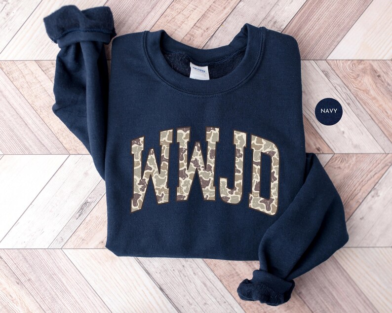 May include: Navy blue sweatshirt with the letters "WWJD" in a camouflage pattern across the chest. The sweatshirt has a crew neck and long sleeves with ribbed cuffs. A small navy circle with the word "NAVY" is in the upper right corner.
