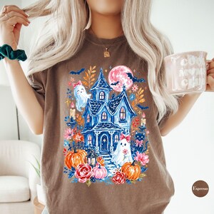 K&ouml;nnte beinhalten: Ein braunes T-Shirt mit einem farbenfrohen Grafikdruck eines blauen Spukhauses, zwei Geistern mit rosa Schleifen, orangefarbenen K&uuml;rbissen, einer schwarzen Katze, einem rosa Mond, Flederm&auml;usen und floralen Elementen, Kerzen und einer Laterne.