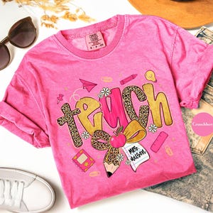 May include: A pink Comfort Colors t-shirt with the word "TEACH" in gold and leopard print, featuring school-themed graphics like an apple, pencil, and paper airplane. The shirt includes the text "Mrs. Wilson".