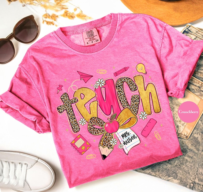May include: A pink Comfort Colors t-shirt with the word "TEACH" in gold and leopard print, featuring school-themed graphics like an apple, pencil, and paper airplane. The shirt includes the text "Mrs. Wilson".