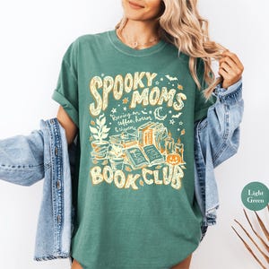 May include: A light green t-shirt featuring a graphic print in yellow and white. The text reads "SPOOKY MOMS RUNNING ON COFFEE, HORROR & CHAPTERS BOOK CLUB". The design includes illustrations of potted plants, stacked books, candles, and a pumpkin.