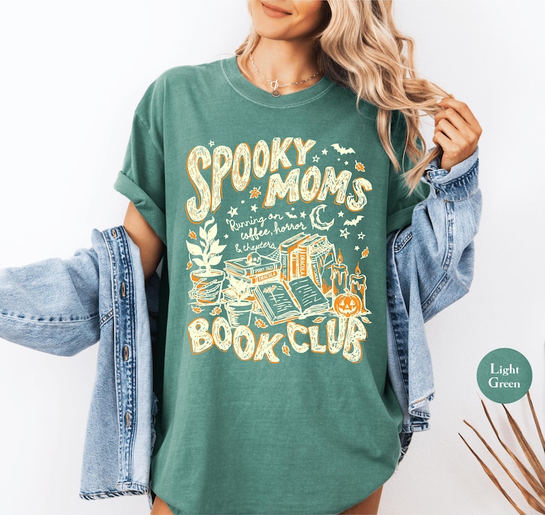 May include: A light green t-shirt featuring a graphic print in yellow and white. The text reads "SPOOKY MOMS RUNNING ON COFFEE, HORROR & CHAPTERS BOOK CLUB". The design includes illustrations of potted plants, stacked books, candles, and a pumpkin.