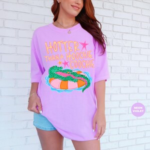 Puede incluir: Camiseta violeta ne&oacute;n con el texto "Hotter Than A Hoochie Coochie" en amarillo y rosa. La camiseta presenta un gr&aacute;fico de un caim&aacute;n verde sobre un flotador inflable naranja y rosa. Corte holgado.