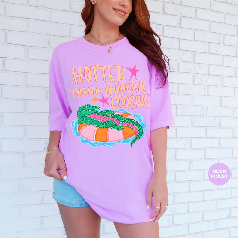 Puede incluir: Camiseta violeta ne&oacute;n con el texto "Hotter Than A Hoochie Coochie" en amarillo y rosa. La camiseta presenta un gr&aacute;fico de un caim&aacute;n verde sobre un flotador inflable naranja y rosa. Corte holgado.