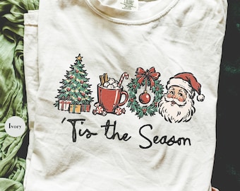 May include: An ivory short-sleeve t-shirt featuring a graphic print of a Christmas tree, a mug of hot cocoa, a wreath, and Santa Claus. Below the illustrations, the text reads 'Tis the Season in a black, handwritten-style script font.