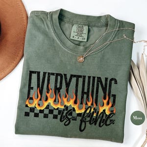 Może przedstawiać: Zielony T-shirt z napisem "Everything is fine" w kolorze czarnym z pomarańczowymi płomieniami i kratką pod spodem. Koszulka wykonana jest z miękkiego, wygodnego materiału.