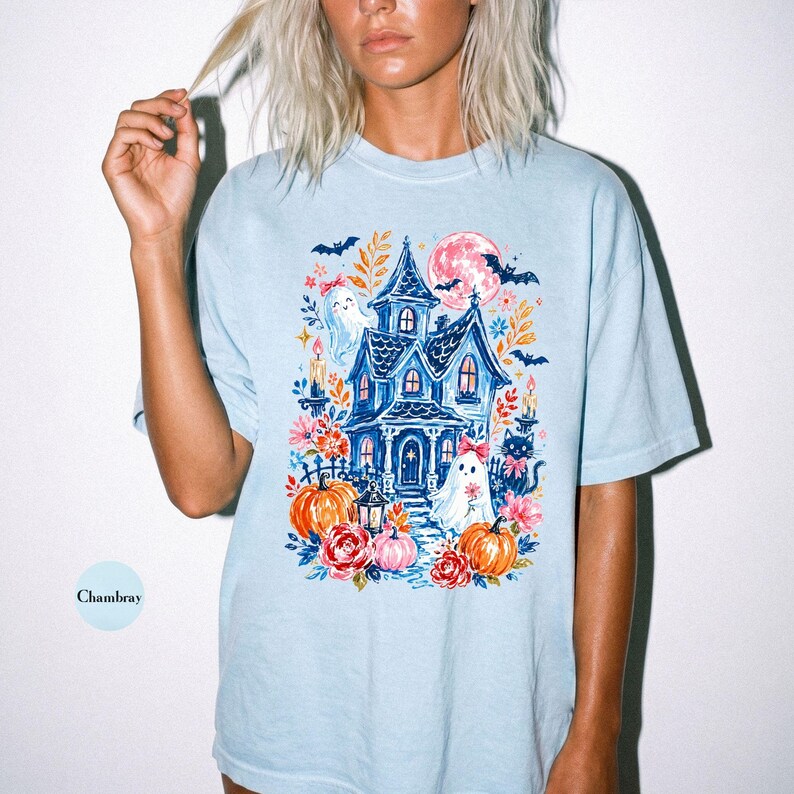 K&ouml;nnte beinhalten: Ein hellblaues T-Shirt mit einem Grafikdruck eines Spukhauses, Geistern, K&uuml;rbissen, Blumen, Flederm&auml;usen und einem Vollmond. Das Design enth&auml;lt eine schwarze Katze, Kerzen und Laub in Orange-, Rosa- und Blaut&ouml;nen. Das Wort Chambray ist aufgedruckt.