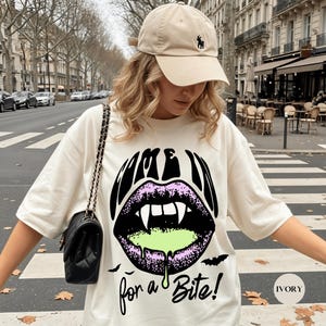 May include: An ivory t-shirt featuring a graphic of black lips with white fangs and green dripping liquid. The text reads "COME IN for a Bite!" with small black bat silhouettes. A beige baseball cap with a small dark logo is worn with the shirt.