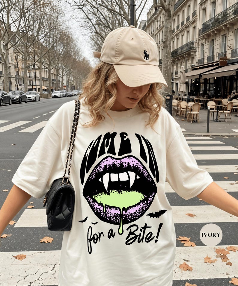 May include: An ivory t-shirt featuring a graphic of black lips with white fangs and green dripping liquid. The text reads "COME IN for a Bite!" with small black bat silhouettes. A beige baseball cap with a small dark logo is worn with the shirt.
