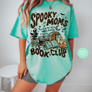 May include: A mint green t-shirt featuring a graphic print with the text "SPOOKY MOMS BOOK CLUB" and "Running on coffee, horror & chapters". The design includes illustrations of stacked books, a potted plant, a pumpkin, candles, and bats.