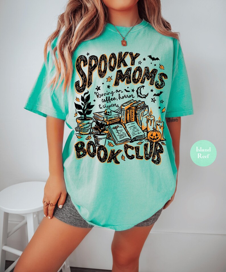 May include: A mint green t-shirt featuring a graphic print with the text "SPOOKY MOMS BOOK CLUB" and "Running on coffee, horror & chapters". The design includes illustrations of stacked books, a potted plant, a pumpkin, candles, and bats.