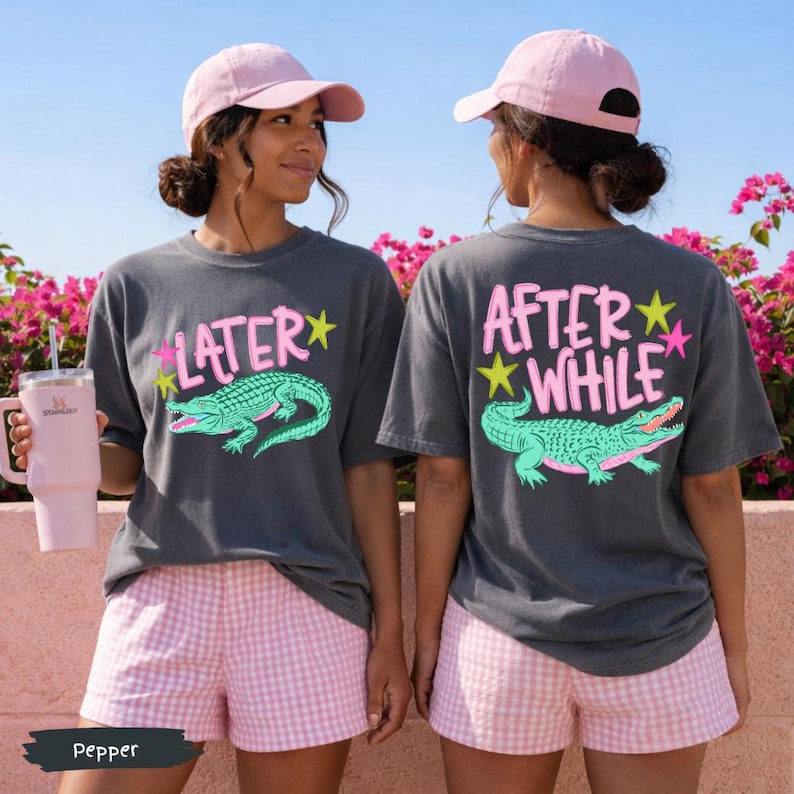 May include: Two women wearing matching gray t-shirts with a cartoon alligator design and the text "LATER" and "AFTER WHILE". They are also wearing pink baseball caps and pink and white checkered shorts. One is holding a pink tumbler.