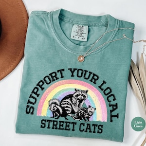 May include: A light green t-shirt featuring the text "Support Your Local Street Cats" in black lettering. The text is encircled by a rainbow with three cartoon animals: a raccoon, a skunk, and an opossum.