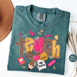 May include: A teal Comfort Colors t-shirt with a "Teach" graphic. The word "Teach" is in a mix of leopard print and pink lettering, with school-themed illustrations like an apple, pencil, and paper airplane. A gold necklace is draped across the shirt.