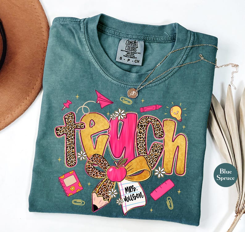 May include: A teal Comfort Colors t-shirt with a "Teach" graphic. The word "Teach" is in a mix of leopard print and pink lettering, with school-themed illustrations like an apple, pencil, and paper airplane. A gold necklace is draped across the shirt.