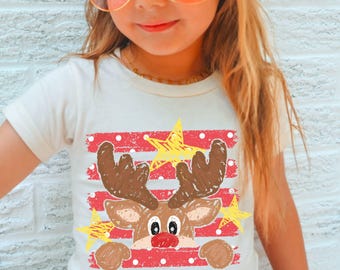 May include: A short-sleeved cream t-shirt featuring a graphic of a brown reindeer with a red nose, positioned behind red horizontal stripes with white polka dots and yellow star illustrations, rendered in a textured, crayon-like artistic style.