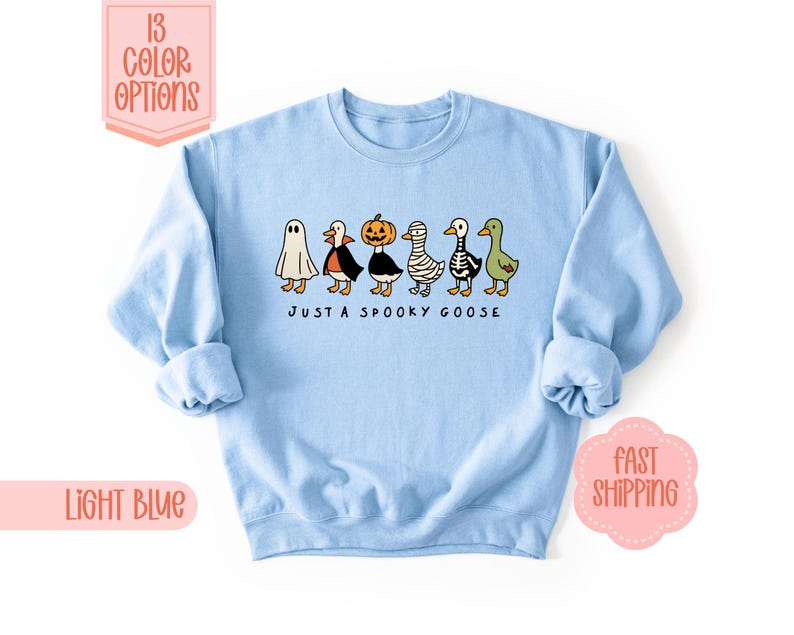 May include: Pale blue sweatshirt with a Halloween-themed design. It features a row of cartoon geese in costumes, including a ghost, vampire, pumpkin, mummy, skeleton, and green goose. The text "JUST A SPOOKY GOOSE" is printed below.
