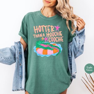 Puede incluir: Camiseta verde claro con el texto "HOTTER THAN A HOOCHIE COOCHIE" en rosa y amarillo. La camiseta presenta un gr&aacute;fico de un caim&aacute;n verde sobre una boya rosa y naranja. La camiseta es de color verde claro.