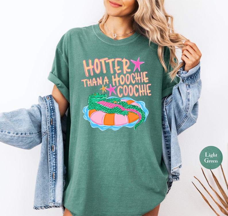 Puede incluir: Camiseta verde claro con el texto "HOTTER THAN A HOOCHIE COOCHIE" en rosa y amarillo. La camiseta presenta un gr&aacute;fico de un caim&aacute;n verde sobre una boya rosa y naranja. La camiseta es de color verde claro.