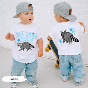 May include: White toddler t-shirt with a raccoon graphic and the text "See Ya Soon" in blue. The back of the shirt also features a raccoon graphic and blue stars. The child is wearing blue jeans and a gray baseball cap.