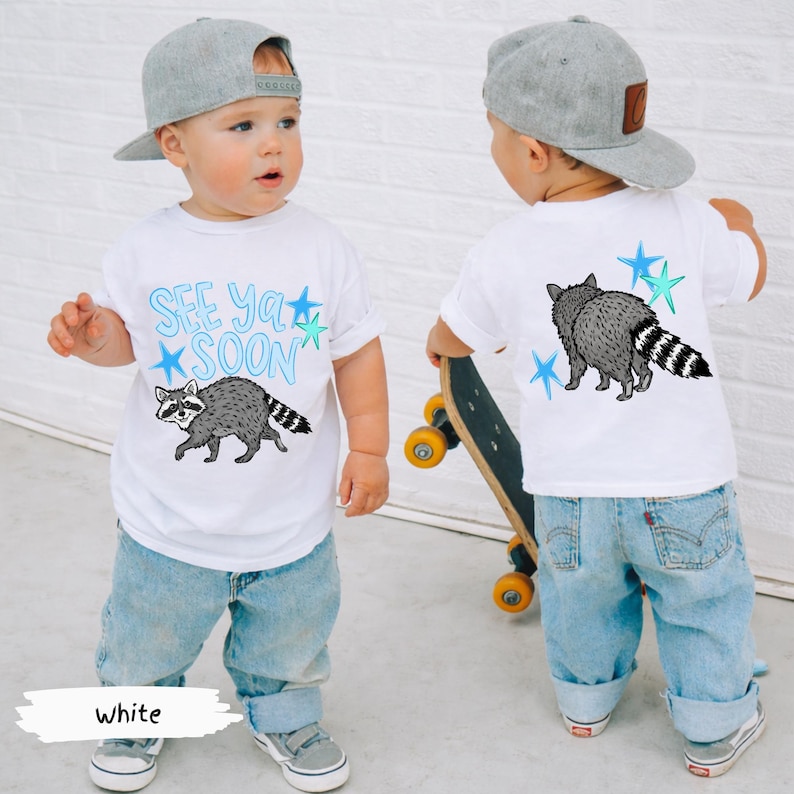 May include: White toddler t-shirt with a raccoon graphic and the text "See Ya Soon" in blue. The back of the shirt also features a raccoon graphic and blue stars. The child is wearing blue jeans and a gray baseball cap.