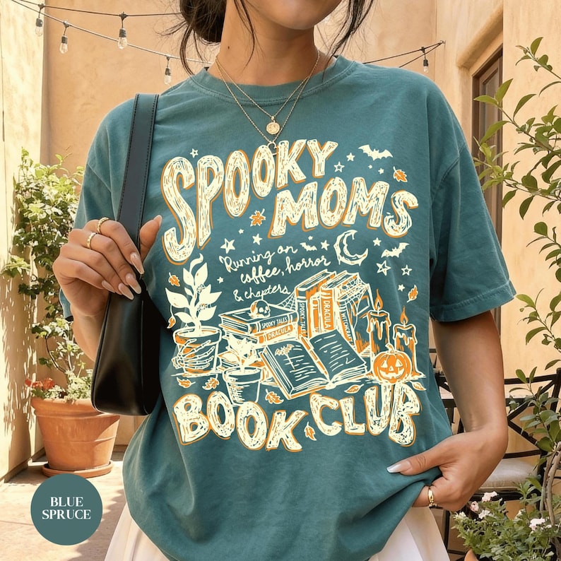 May include: A blue spruce short-sleeve t-shirt featuring a graphic print with the text SPOOKY MOMS RUNNING ON COFFEE, HORROR & CHAPTERS BOOK CLUB. The design includes illustrations of potted plants, stacked books, candles, and a pumpkin.