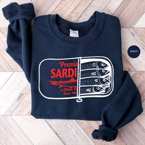 May include: Navy blue sweatshirt featuring a graphic of sardines in a can. The design includes the text "Premium Sardines in Olive Oil Since 1901" in red and white. The sweatshirt is made of a soft material.