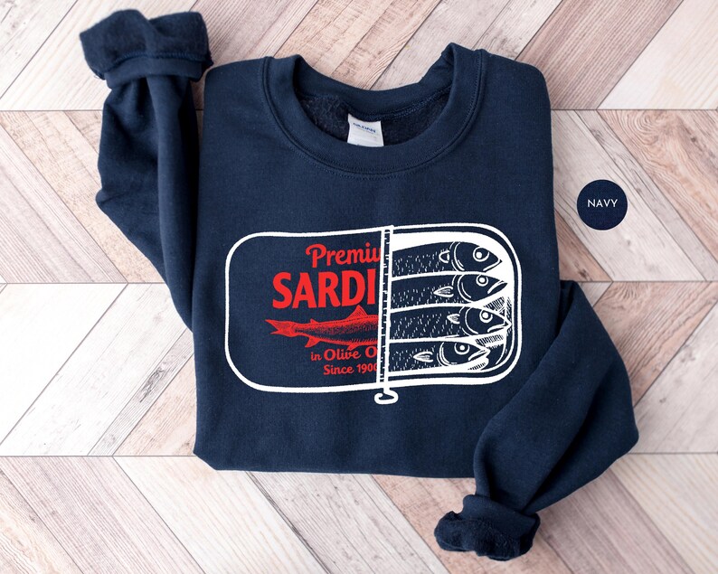 May include: Navy blue sweatshirt featuring a graphic of sardines in a can. The design includes the text "Premium Sardines in Olive Oil Since 1901" in red and white. The sweatshirt is made of a soft material.