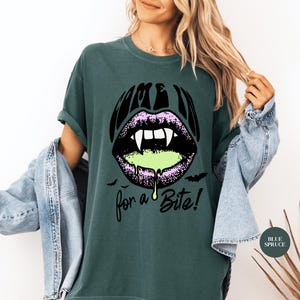 May include: A dark spruce green t-shirt featuring a graphic of black lips with white fangs and dripping neon green liquid. The text reads "COME IN" above the lips and "for a Bite!" below, accompanied by small black bat silhouettes.