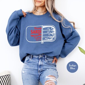 May include: Indigo blue sweatshirt featuring a sardine can graphic. The graphic includes the words "Premier Sardines" in red, with a white outline. The sardine can graphic shows three fish. The sweatshirt is a crew neck style.
