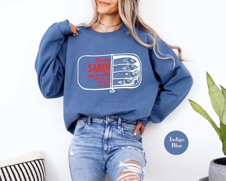 May include: Indigo blue sweatshirt featuring a sardine can graphic. The graphic includes the words "Premier Sardines" in red, with a white outline. The sardine can graphic shows three fish. The sweatshirt is a crew neck style.