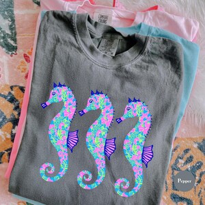 May include: A stack of Comfort Colors t-shirts in gray, pink, and blue. The gray shirt features three seahorse designs in pink, blue, and green. The shirts are folded and stacked on a patterned surface.
