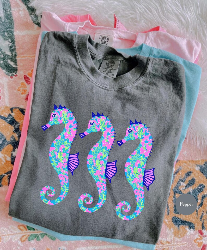 May include: A stack of Comfort Colors t-shirts in gray, pink, and blue. The gray shirt features three seahorse designs in pink, blue, and green. The shirts are folded and stacked on a patterned surface.