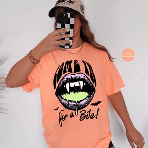 May include: A neon cantaloupe orange short-sleeve t-shirt featuring a graphic of black lips with white fangs and dripping lime green liquid. The text reads "COME IN for a Bite!" in a black stylized font, accompanied by two small black bat silhouettes.