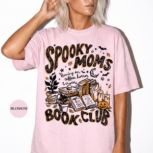 May include: A light pink t-shirt featuring a graphic print with the text "SPOOKY MOMS BOOK CLUB" and "Running on coffee, horror & chapters". The design includes illustrations of stacked books, a skull, a pumpkin, candles, and a potted plant.