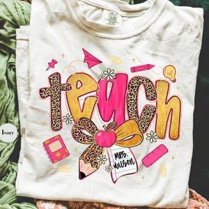 May include: Ivory-colored t-shirt with the word "TEACH" in gold, pink, and leopard print. The design includes an apple, pencil, notebook, and other school-themed graphics. The shirt also has the text "Mrs. Wilson".