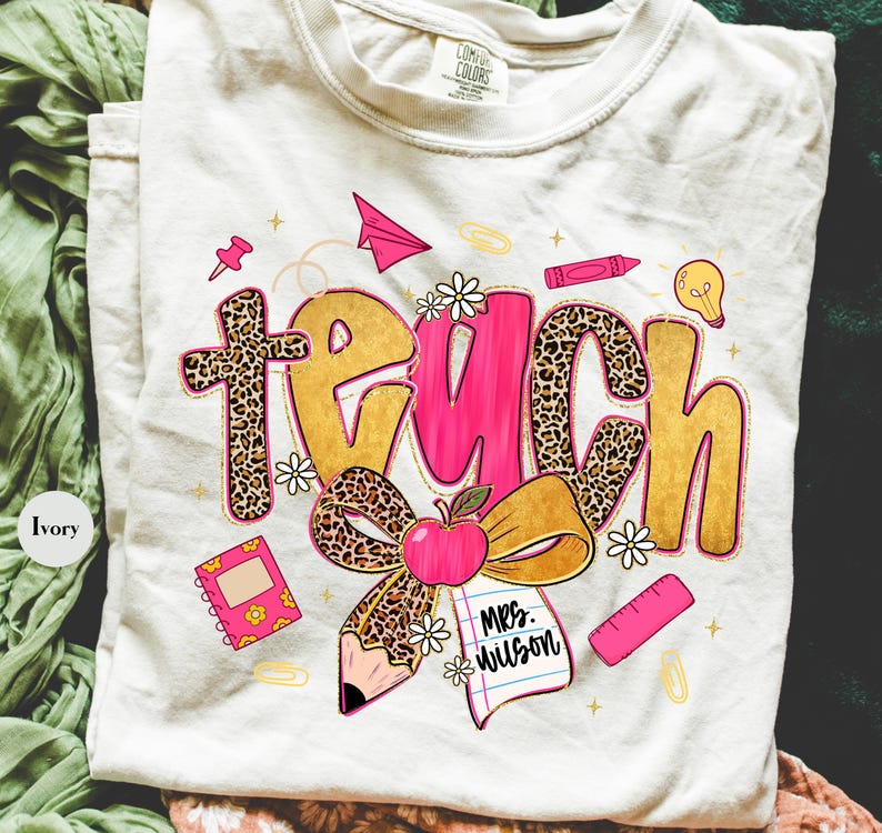 May include: Ivory-colored t-shirt with the word "TEACH" in gold, pink, and leopard print. The design includes an apple, pencil, notebook, and other school-themed graphics. The shirt also has the text "Mrs. Wilson".