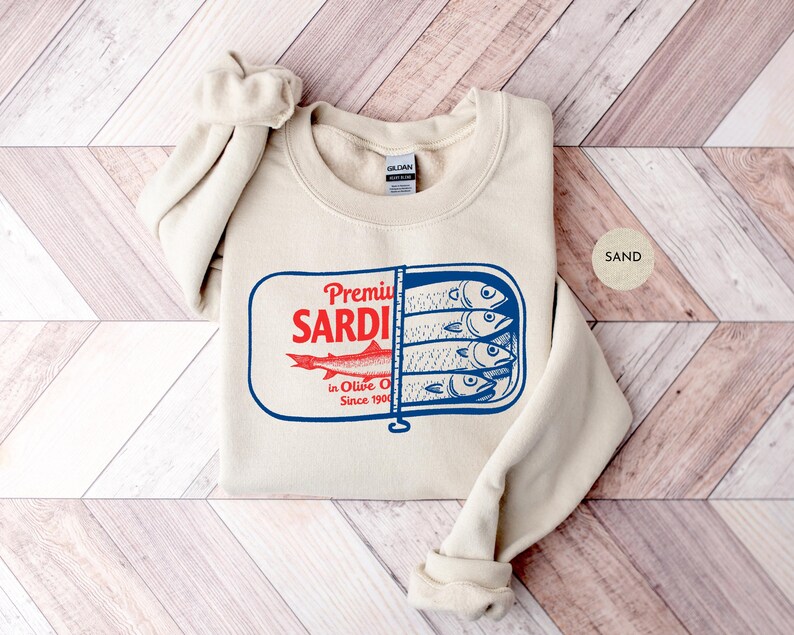 May include: Sand-colored sweatshirt featuring a graphic of a sardine can with the words "Premium Sardi" in red and blue. The design includes illustrations of sardines and the text "in Olive Oil Since 1900".