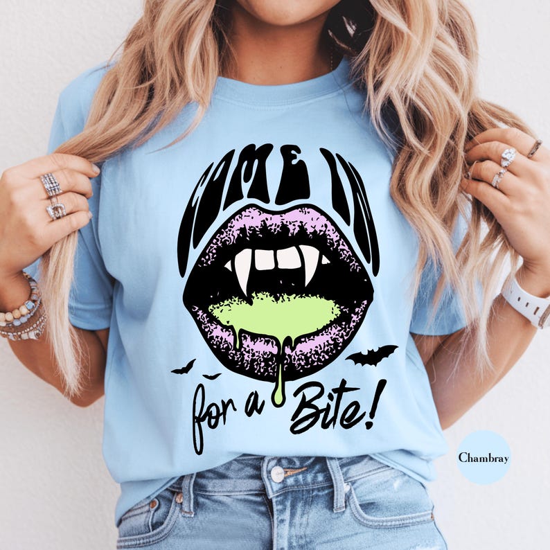 May include: A light blue t-shirt featuring a graphic of black lips with white fangs and green slime dripping from the mouth. The text "COME IN" is printed above the lips and "for a Bite!" below, accompanied by small black bat silhouettes.