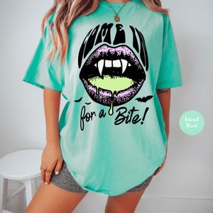 May include: A mint green t-shirt featuring a graphic of black lips with white vampire fangs and dripping neon green liquid. The text reads "COME IN" above the lips and "for a Bite!" below, accompanied by two small black bat silhouettes.