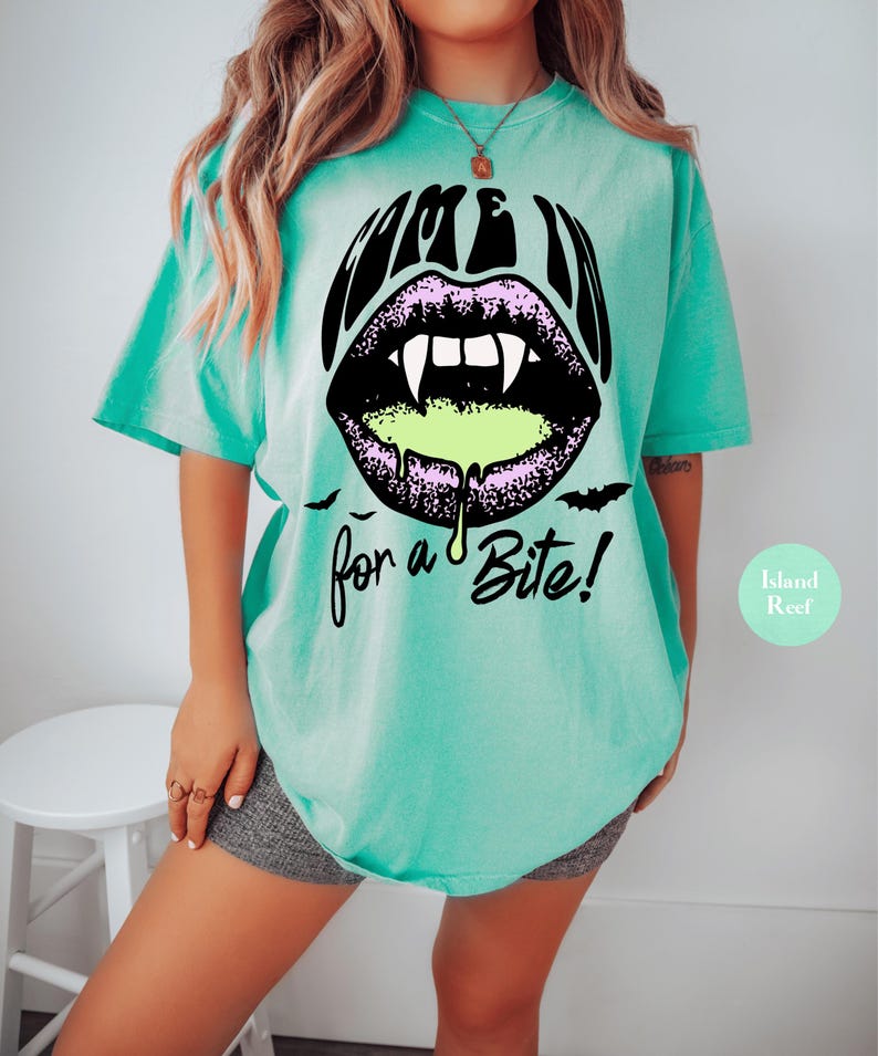 May include: A mint green t-shirt featuring a graphic of black lips with white vampire fangs and dripping neon green liquid. The text reads "COME IN" above the lips and "for a Bite!" below, accompanied by two small black bat silhouettes.